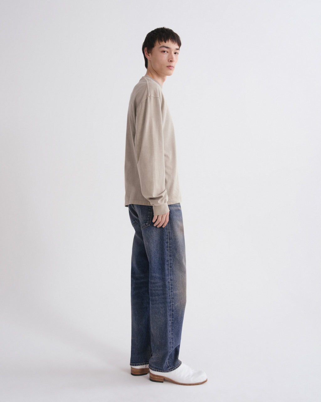 A model standing in profile wearing a light beige knit sweater, wide-leg blue jeans, and white leather loafers against a plain light gray studio background.