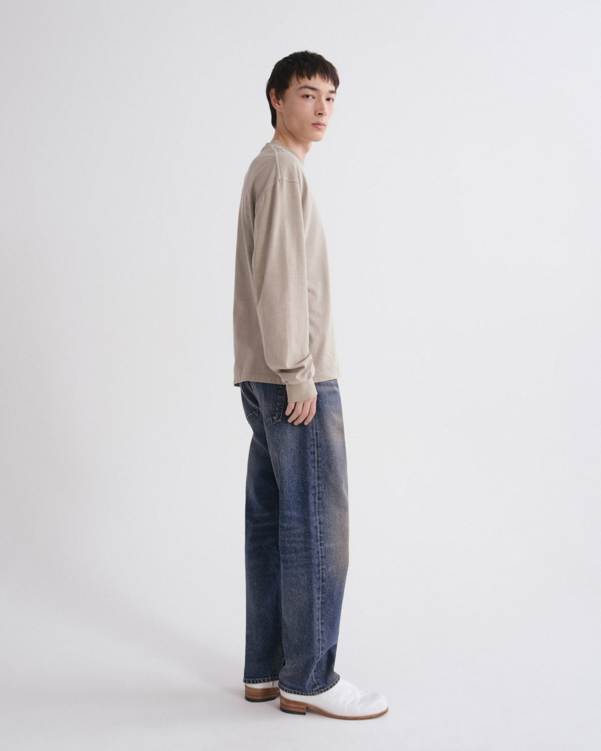 A model standing in profile wearing a light beige knit sweater, wide-leg blue jeans, and white leather loafers against a plain light gray studio background.