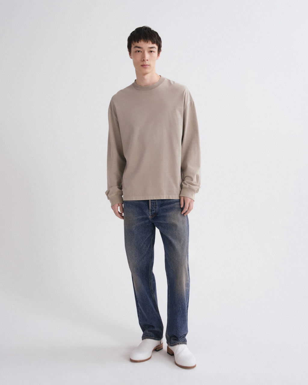 A man modeling a light beige long-sleeve crewneck sweater, blue jeans, and white shoes against a plain light gray studio background.