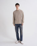 A man modeling a light beige long-sleeve crewneck sweater, blue jeans, and white shoes against a plain light gray studio background.