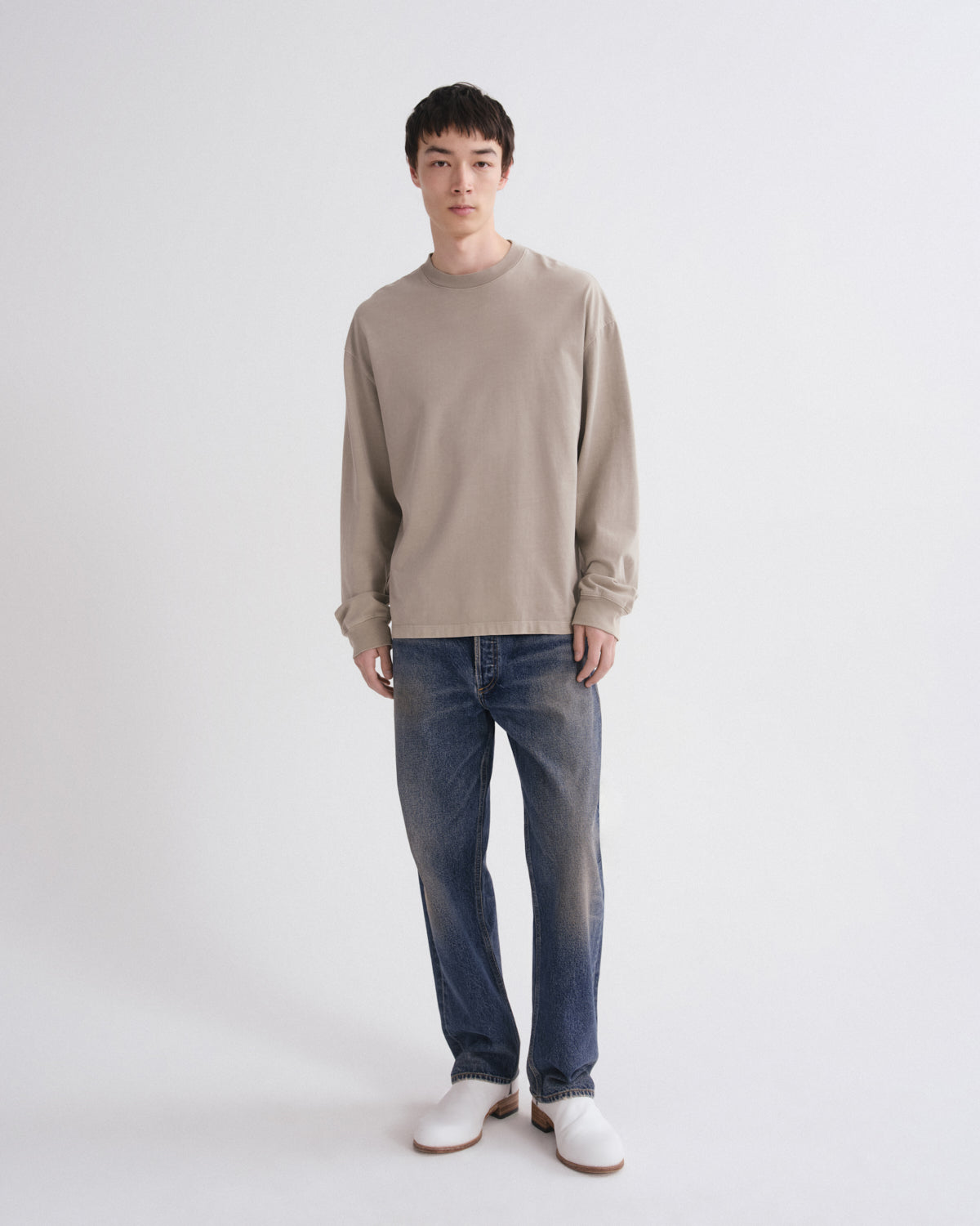 A man modeling a light beige long-sleeve crewneck sweater, blue jeans, and white shoes against a plain light gray studio background.