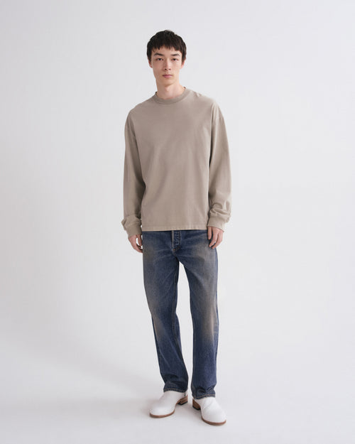 A man modeling a light beige long-sleeve crewneck sweater, blue jeans, and white shoes against a plain light gray studio background.