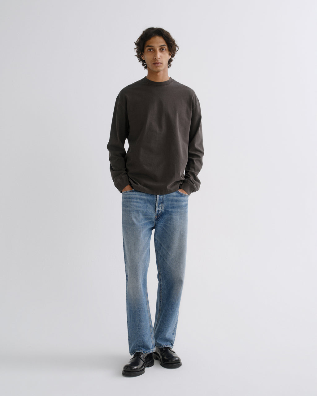 A man wearing a dark charcoal gray long-sleeve crewneck sweatshirt, light blue straight-leg jeans, and black leather shoes, standing against a light gray studio background.