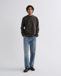 A man wearing a dark charcoal gray long-sleeve crewneck sweatshirt, light blue straight-leg jeans, and black leather shoes, standing against a light gray studio background.