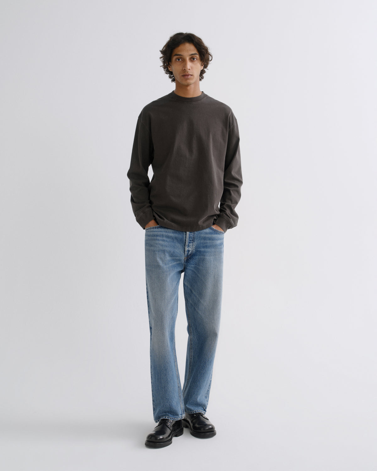 A man wearing a dark charcoal gray long-sleeve crewneck sweatshirt, light blue straight-leg jeans, and black leather shoes, standing against a light gray studio background.