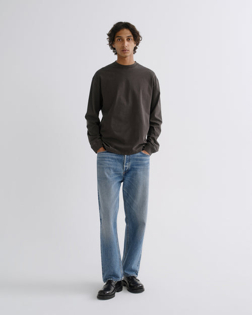 A man wearing a dark charcoal gray long-sleeve crewneck sweatshirt, light blue straight-leg jeans, and black leather shoes, standing against a light gray studio background.