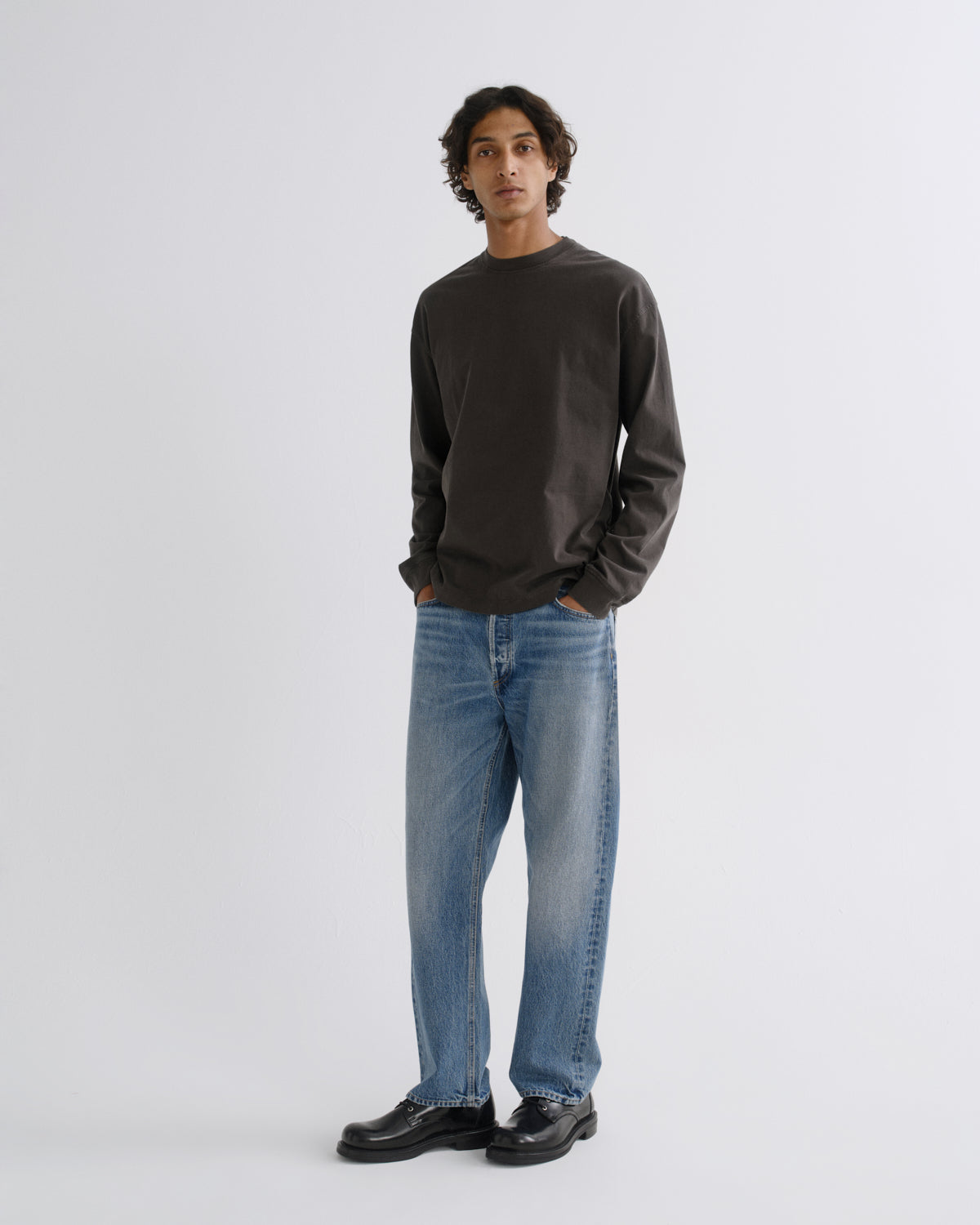 A man modeling a dark brown long-sleeve crewneck sweater, blue jeans, and black leather shoes against a light gray studio background.