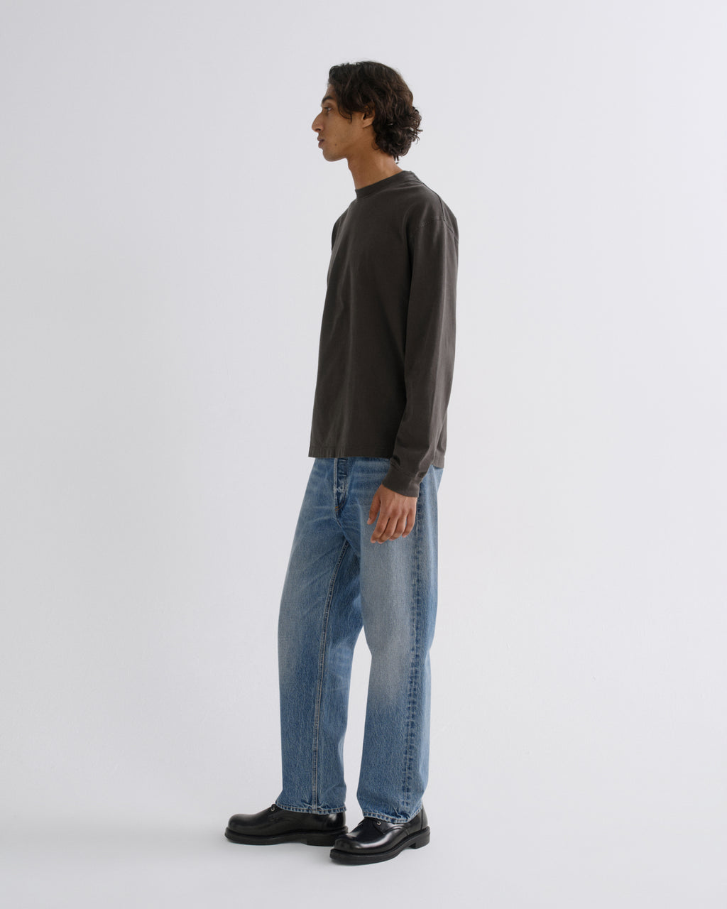 A man in profile wearing a dark charcoal long-sleeve shirt, light blue jeans, and black shoes, standing against a plain light gray studio background.