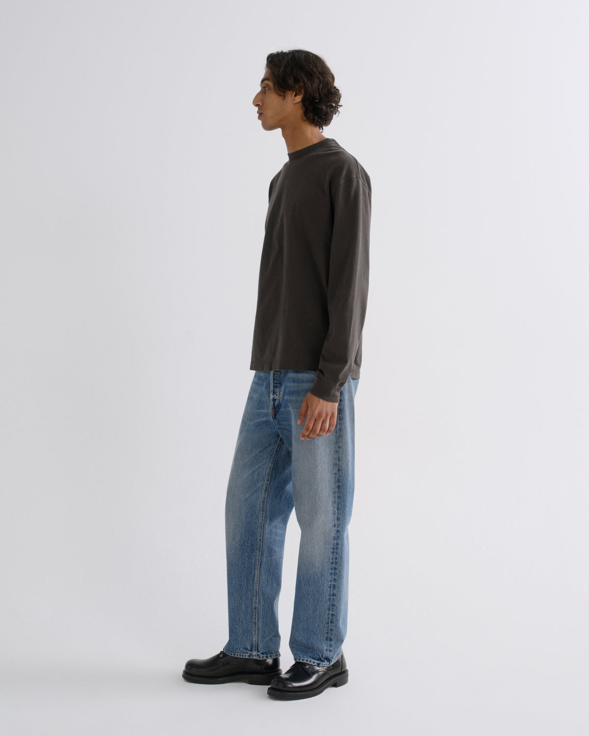 A man in profile wearing a dark charcoal long-sleeve shirt, light blue jeans, and black shoes, standing against a plain light gray studio background.
