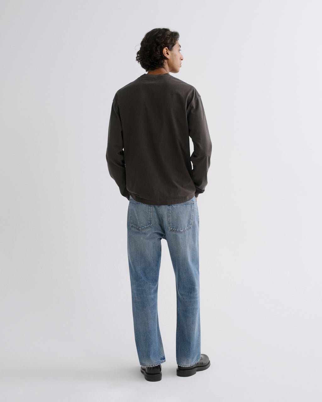 A man viewed from behind wearing a dark charcoal long-sleeve crewneck shirt, light blue jeans, and black shoes, standing against a light gray studio background.