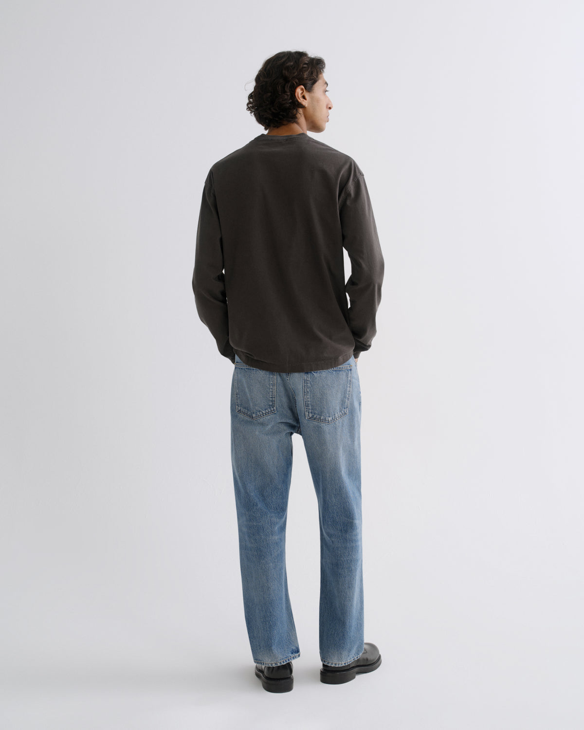 A man viewed from behind wearing a dark charcoal long-sleeve crewneck shirt, light blue jeans, and black shoes, standing against a light gray studio background.