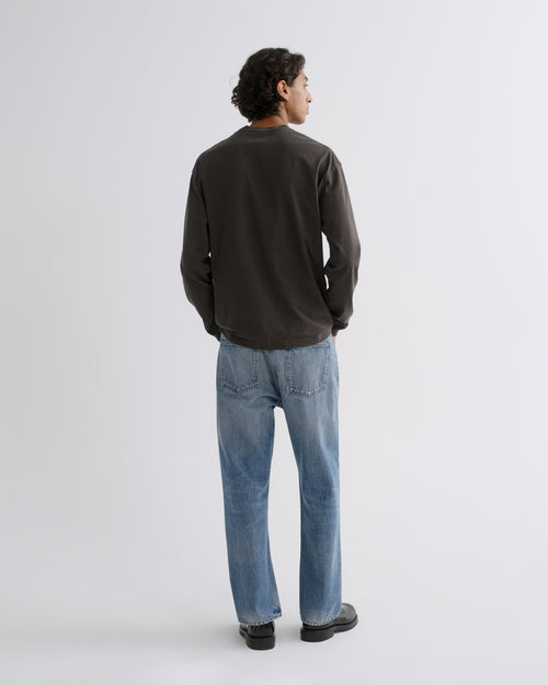 A man viewed from behind wearing a dark charcoal long-sleeve crewneck shirt, light blue jeans, and black shoes, standing against a light gray studio background.