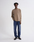 A man wearing a beige long-sleeve crewneck shirt with a chest pocket, blue jeans, and black shoes, standing against a plain white studio background.
