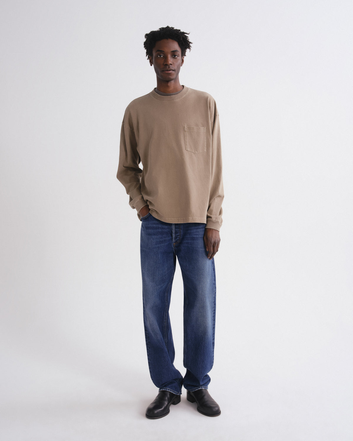 A man wearing a beige long-sleeve crewneck shirt with a chest pocket, blue jeans, and black shoes, standing against a plain white studio background.