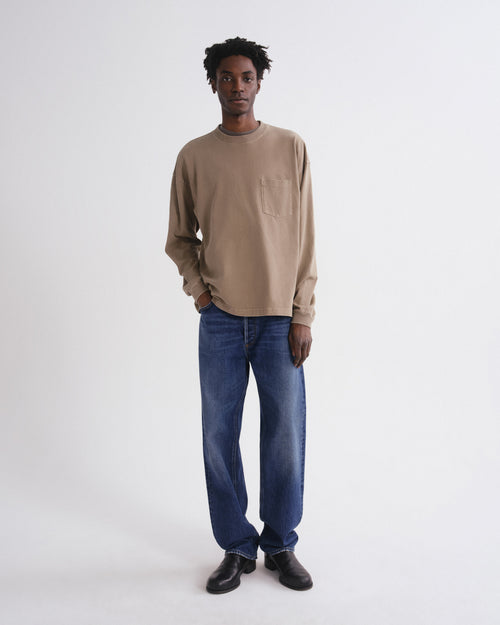 A man wearing a beige long-sleeve crewneck shirt with a chest pocket, blue jeans, and black shoes, standing against a plain white studio background.