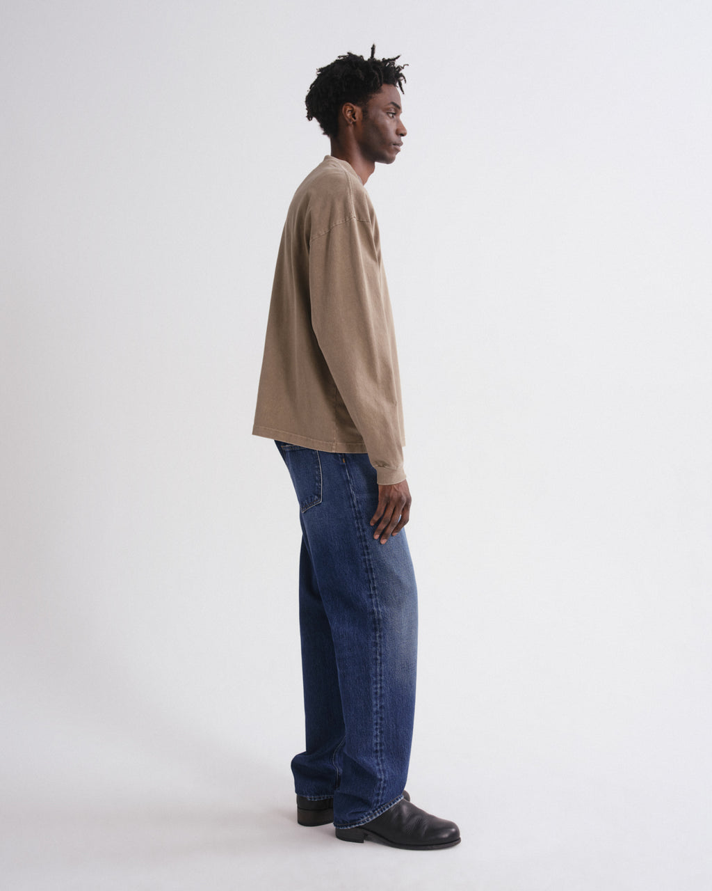 A man in a side profile wearing a beige long-sleeve crewneck sweatshirt, blue jeans, and black leather shoes, standing against a plain white studio background.