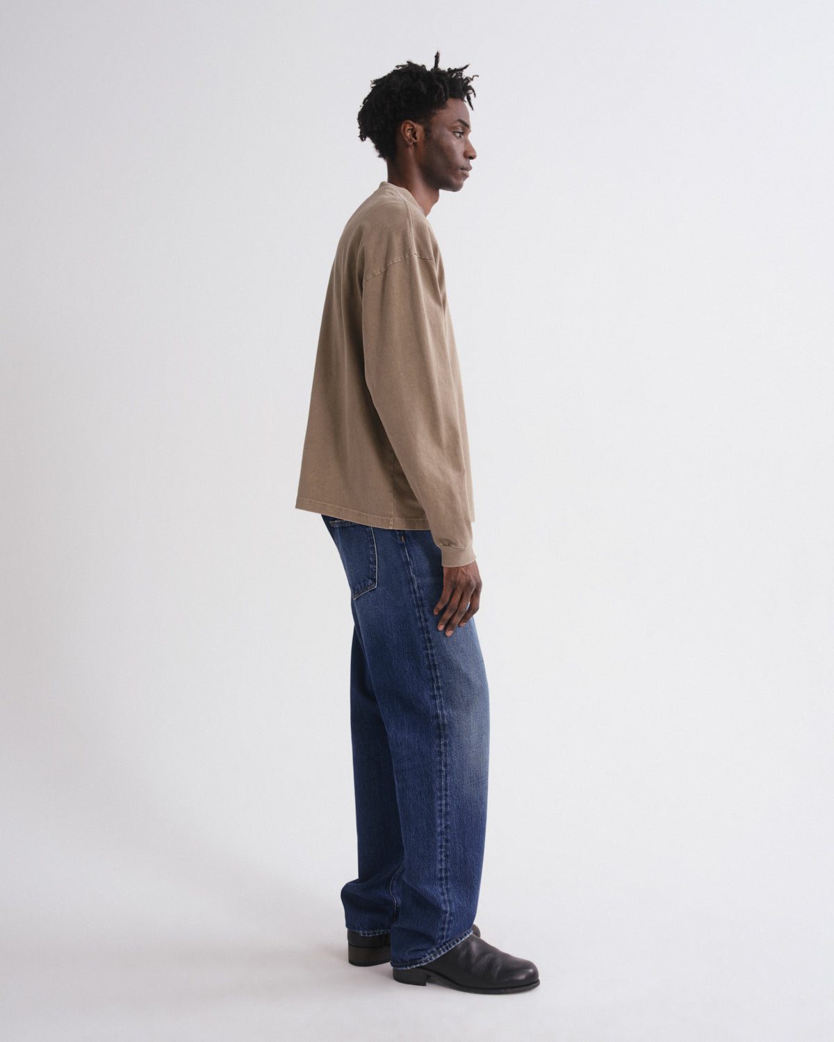 A man in a side profile wearing a beige long-sleeve crewneck sweatshirt, blue jeans, and black leather shoes, standing against a plain white studio background.