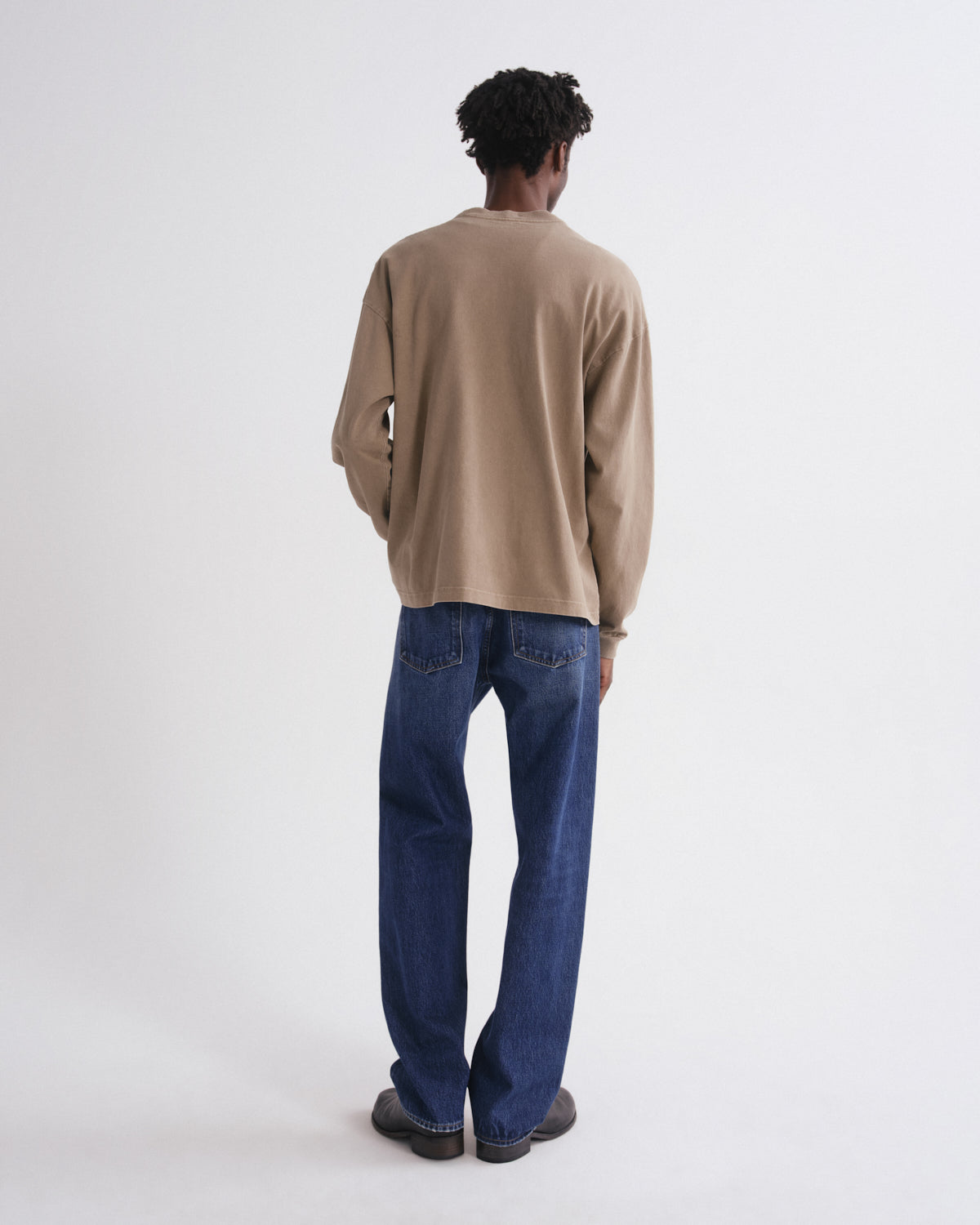 A man viewed from behind wearing a beige long-sleeve knit sweater and dark blue straight-leg jeans against a plain white studio background.