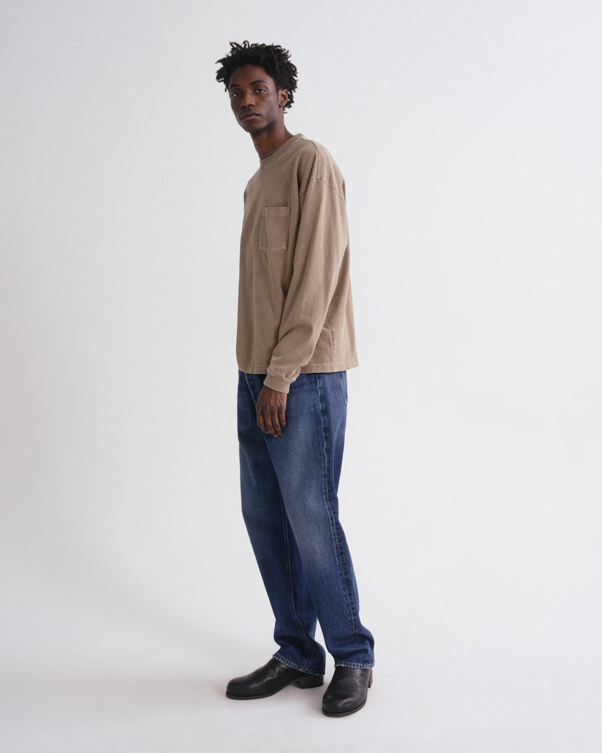 A model wearing a beige long-sleeve crewneck shirt, blue jeans, and black leather shoes, standing in profile against a plain light gray studio background.
