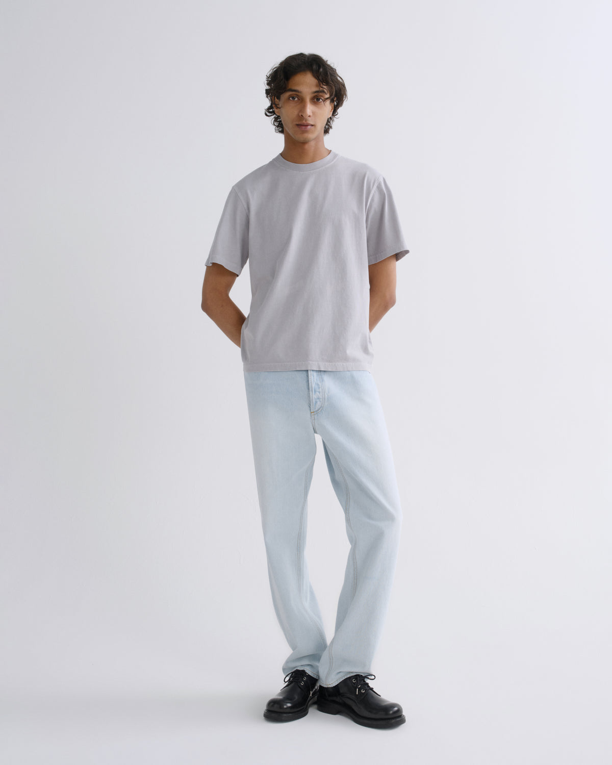 A man modeling a light gray crewneck t-shirt and light blue jeans with black shoes, standing against a plain light gray studio background.