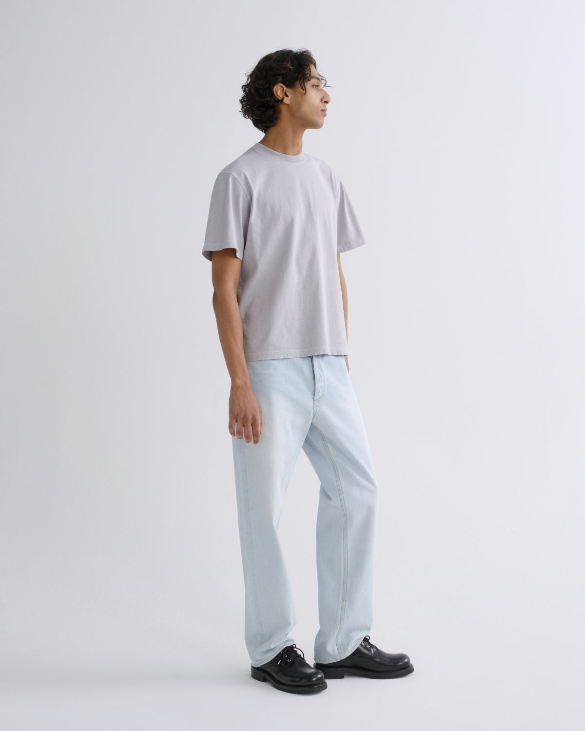 A man wearing a light gray crewneck t-shirt, light blue relaxed-fit jeans, and black leather dress shoes, standing in profile against a solid light gray studio background.