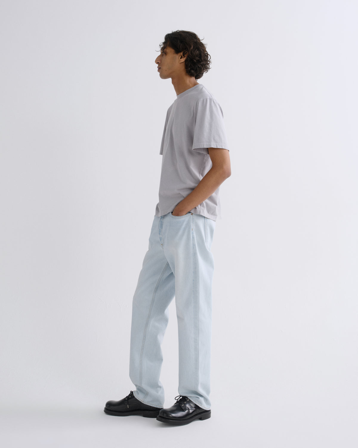 A man in a light gray t-shirt and light blue relaxed-fit trousers, standing in profile against a plain white studio background.