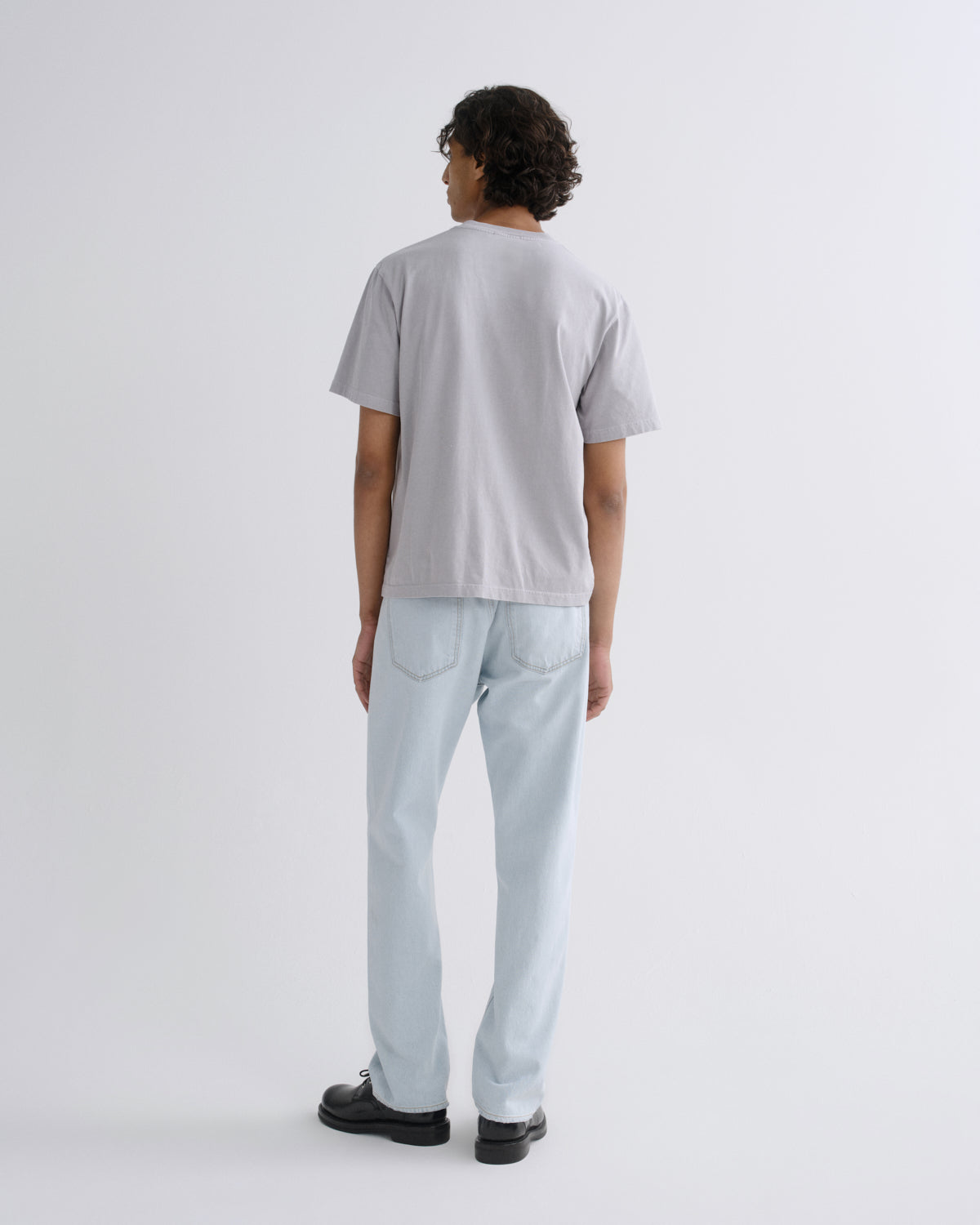 A man is shown from behind wearing a light gray short-sleeve t-shirt, light-wash blue jeans, and black shoes, standing against a plain light gray studio background.