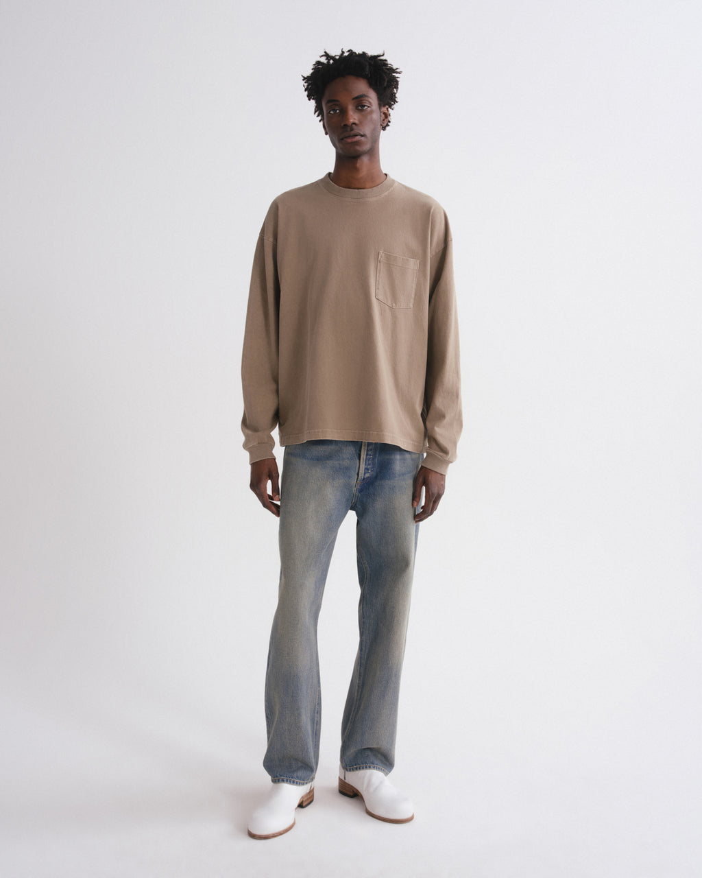 A model stands against a plain light gray studio background wearing a beige long-sleeve crewneck sweatshirt, light-wash blue jeans, and white shoes.