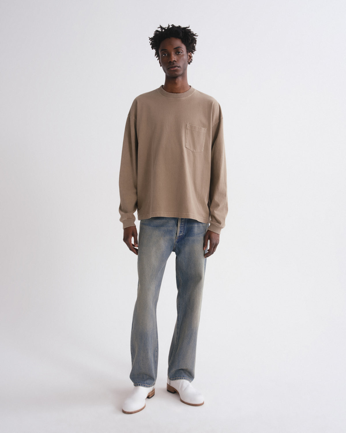 A model stands against a plain light gray studio background wearing a beige long-sleeve crewneck sweatshirt, light-wash blue jeans, and white shoes.
