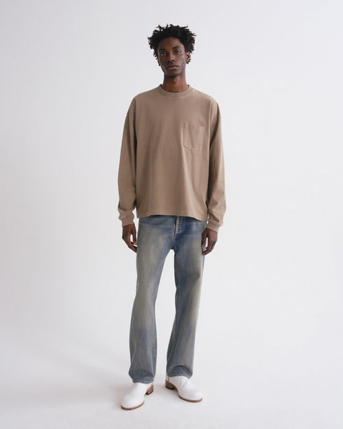 A model stands against a plain light gray studio background wearing a beige long-sleeve crewneck sweatshirt, light-wash blue jeans, and white shoes.