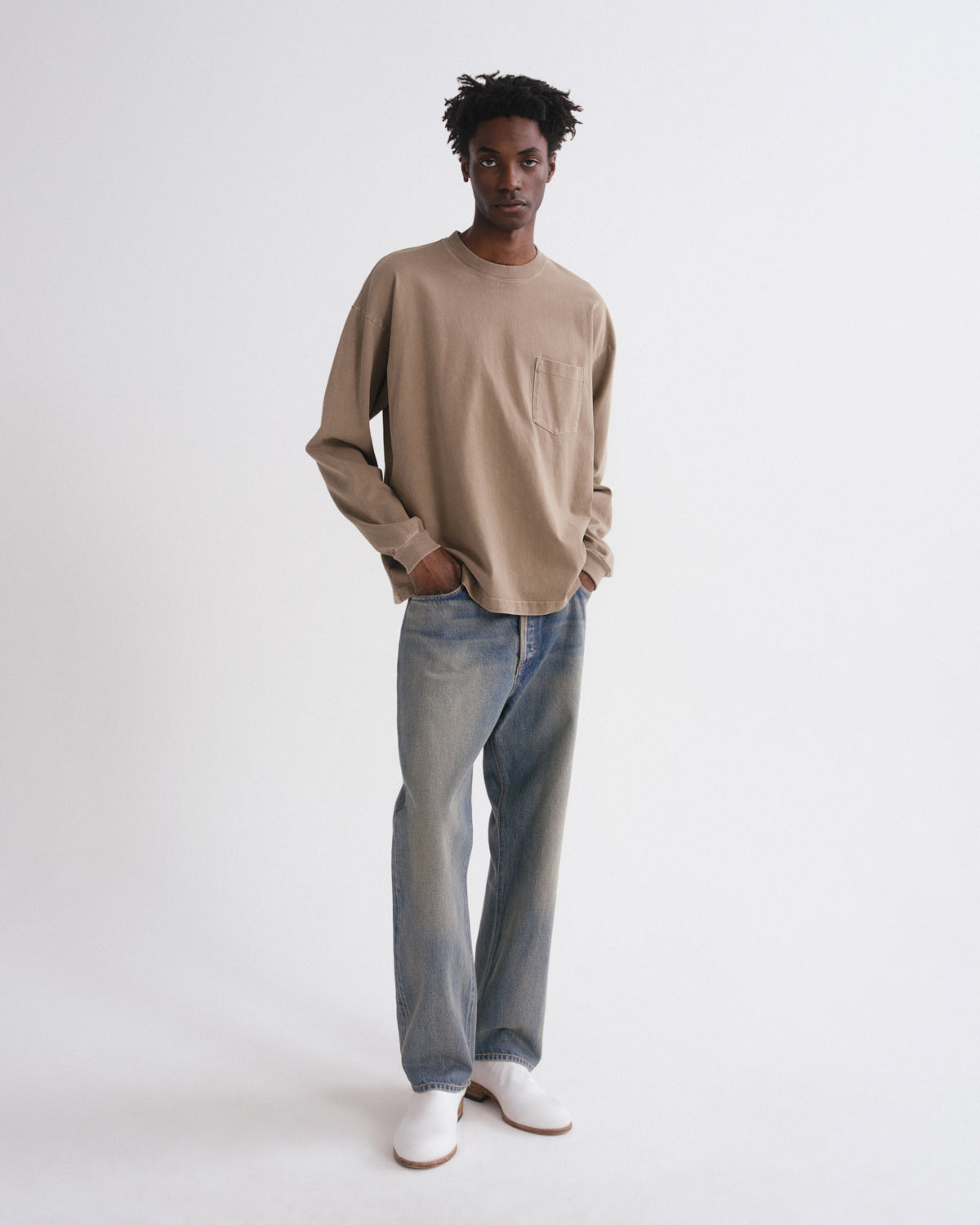 A man modeling a beige long-sleeve crewneck shirt with a chest pocket, paired with light blue jeans and white slip-on shoes, standing against a plain white studio background.