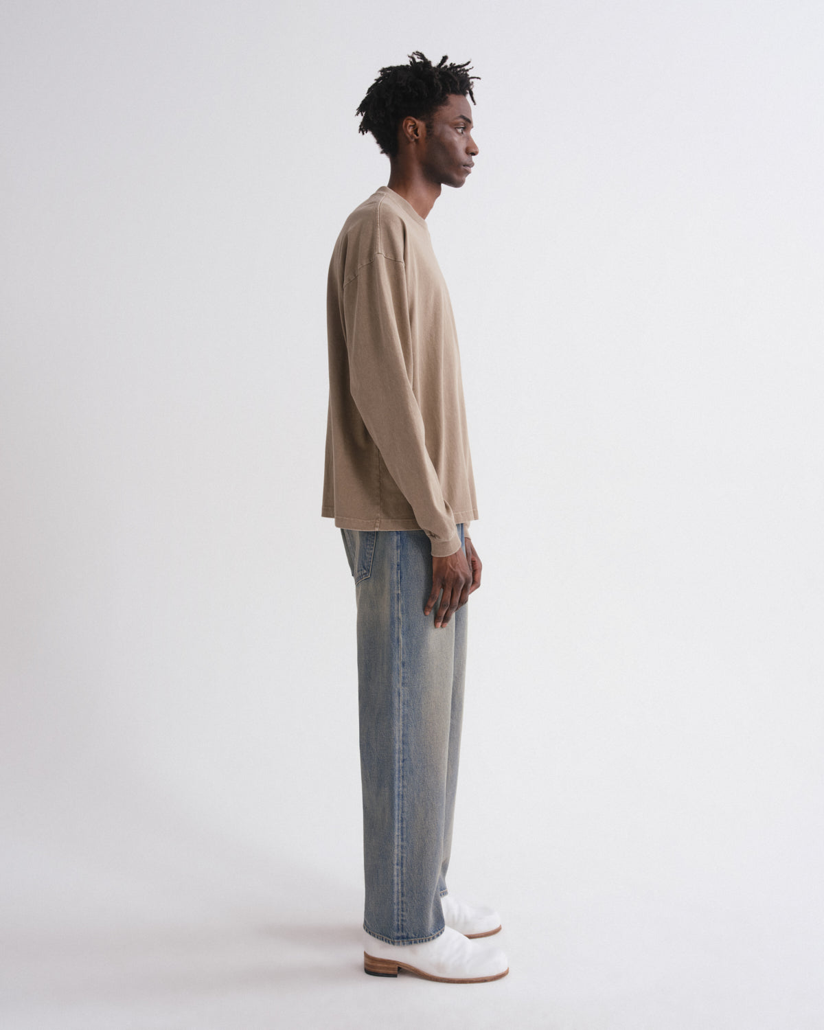 A man stands in profile wearing a beige long-sleeve knit top, light blue wide-leg jeans, and white shoes against a plain light gray studio background.