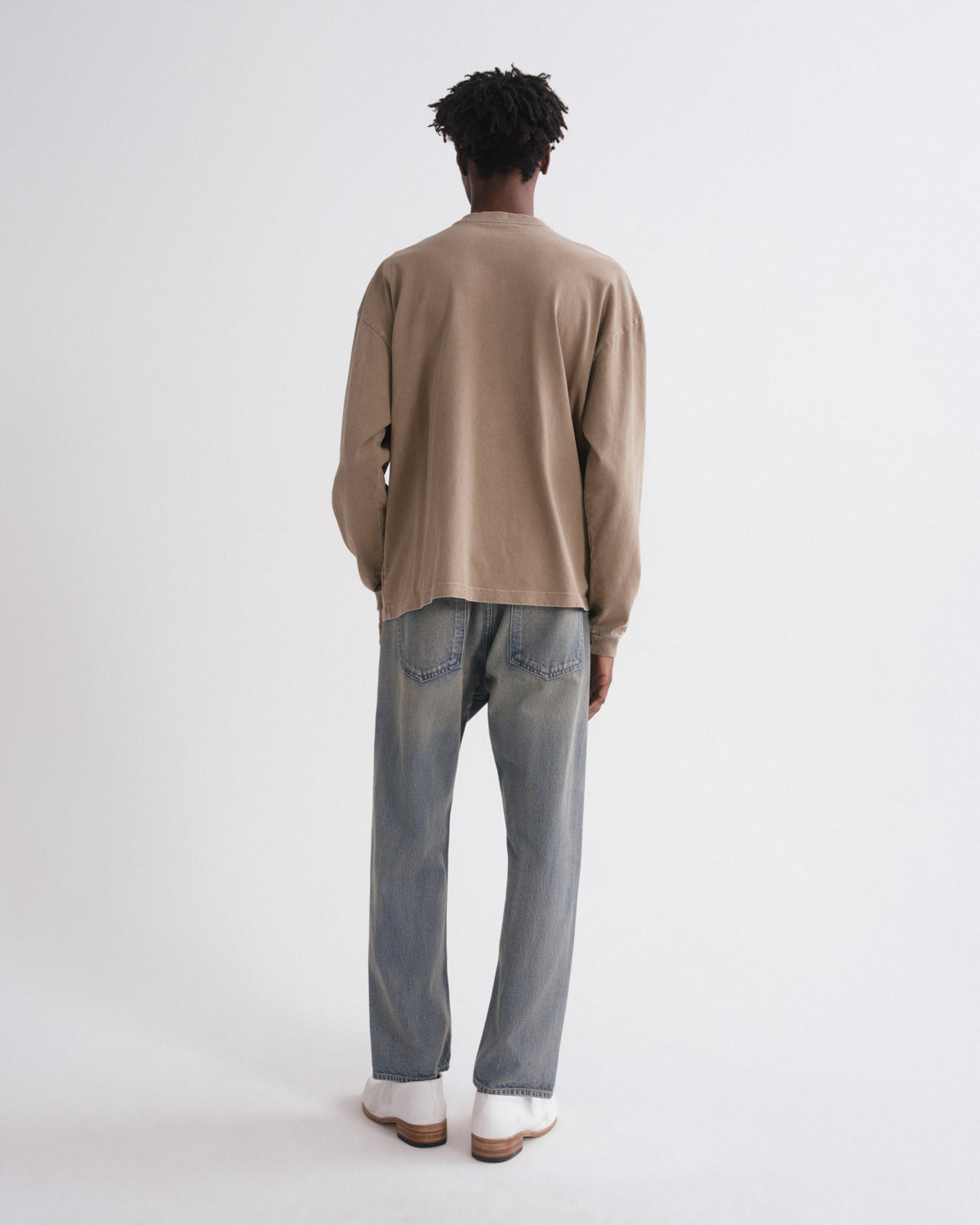 A model viewed from behind wearing a beige long-sleeve knit top, light-wash blue jeans, and white shoes, standing against a plain white studio background.