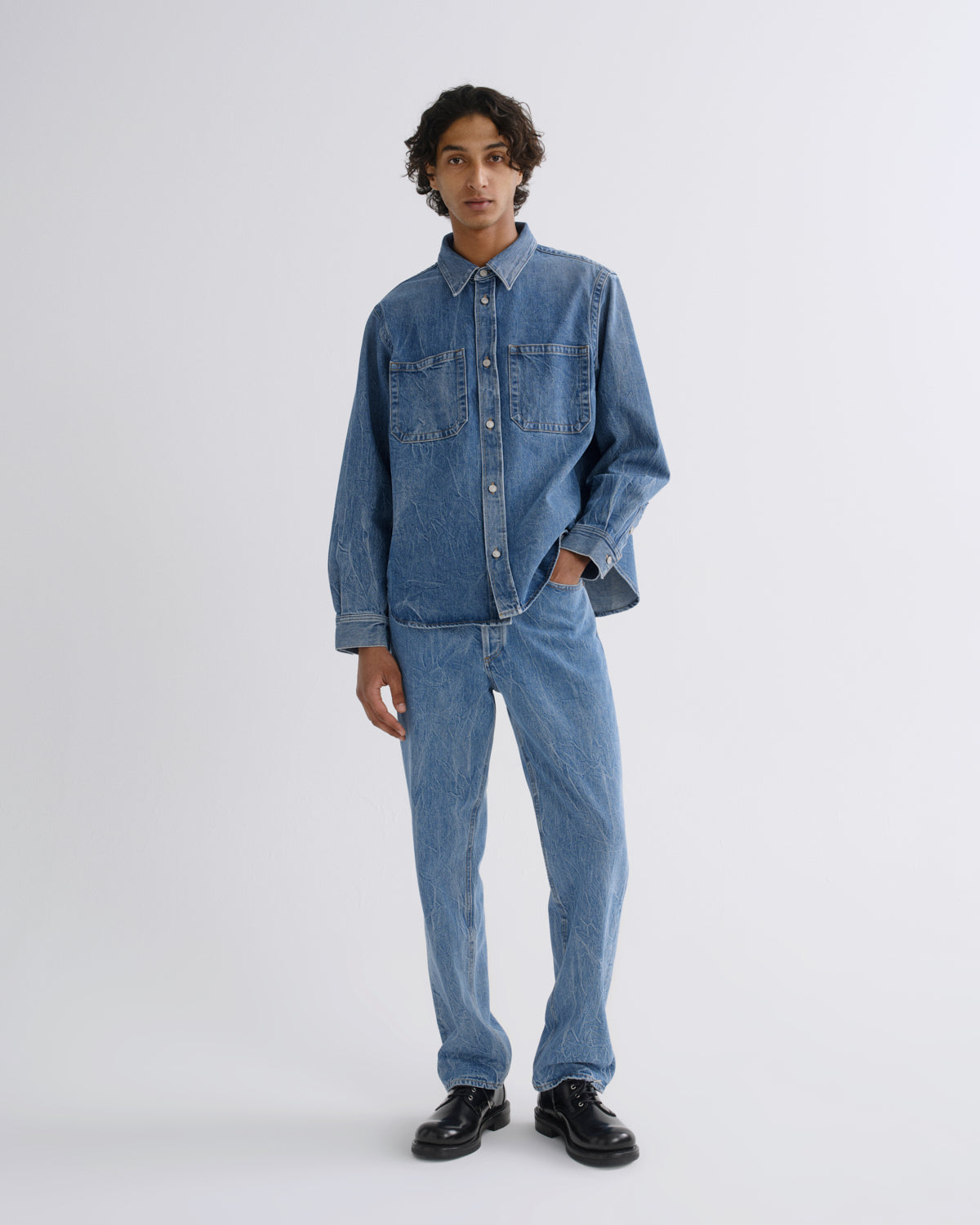 A model stands against a light gray studio background wearing a matching blue denim set consisting of a long-sleeve button-up shirt and straight-leg jeans, paired with black shoes.