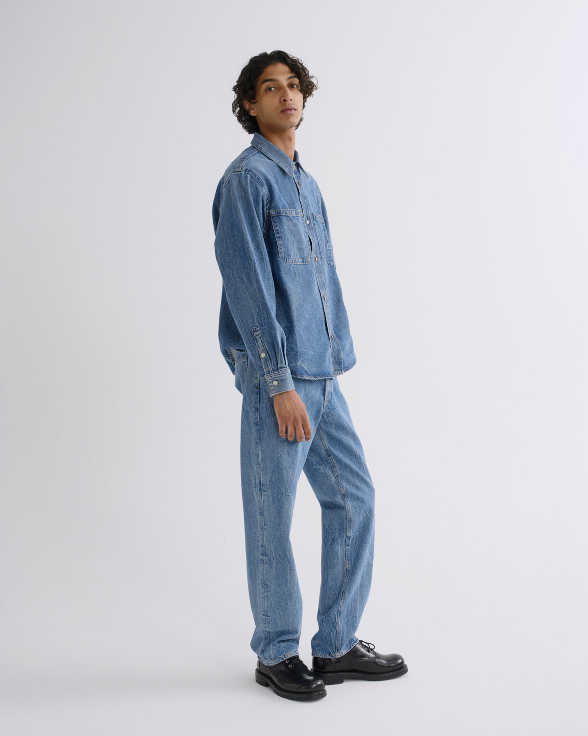A man wearing a light blue denim long-sleeve button-up shirt and matching light blue denim jeans, paired with black leather shoes, standing in profile against a light gray studio background.