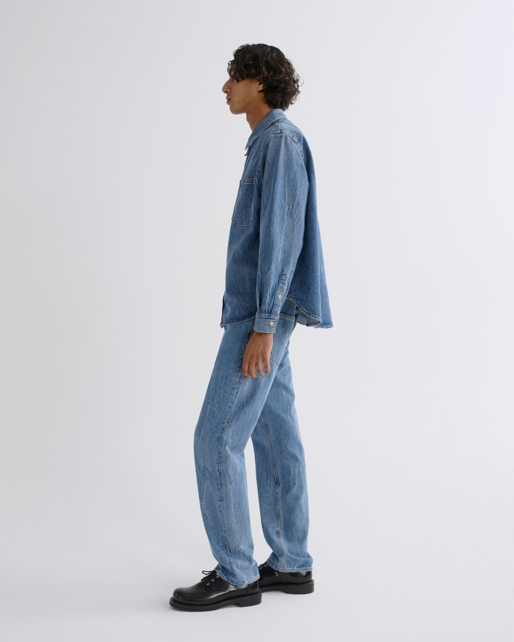 A model shown in profile wearing a light blue denim jacket and matching light blue denim jeans, paired with black leather shoes, against a plain light gray studio background.