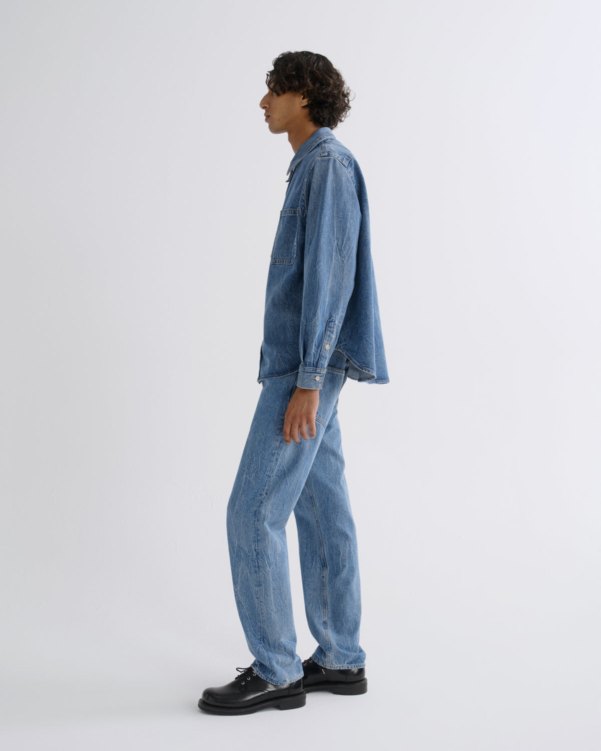 A model shown in profile wearing a light blue denim jacket and matching light blue denim jeans, paired with black leather shoes, against a plain light gray studio background.
