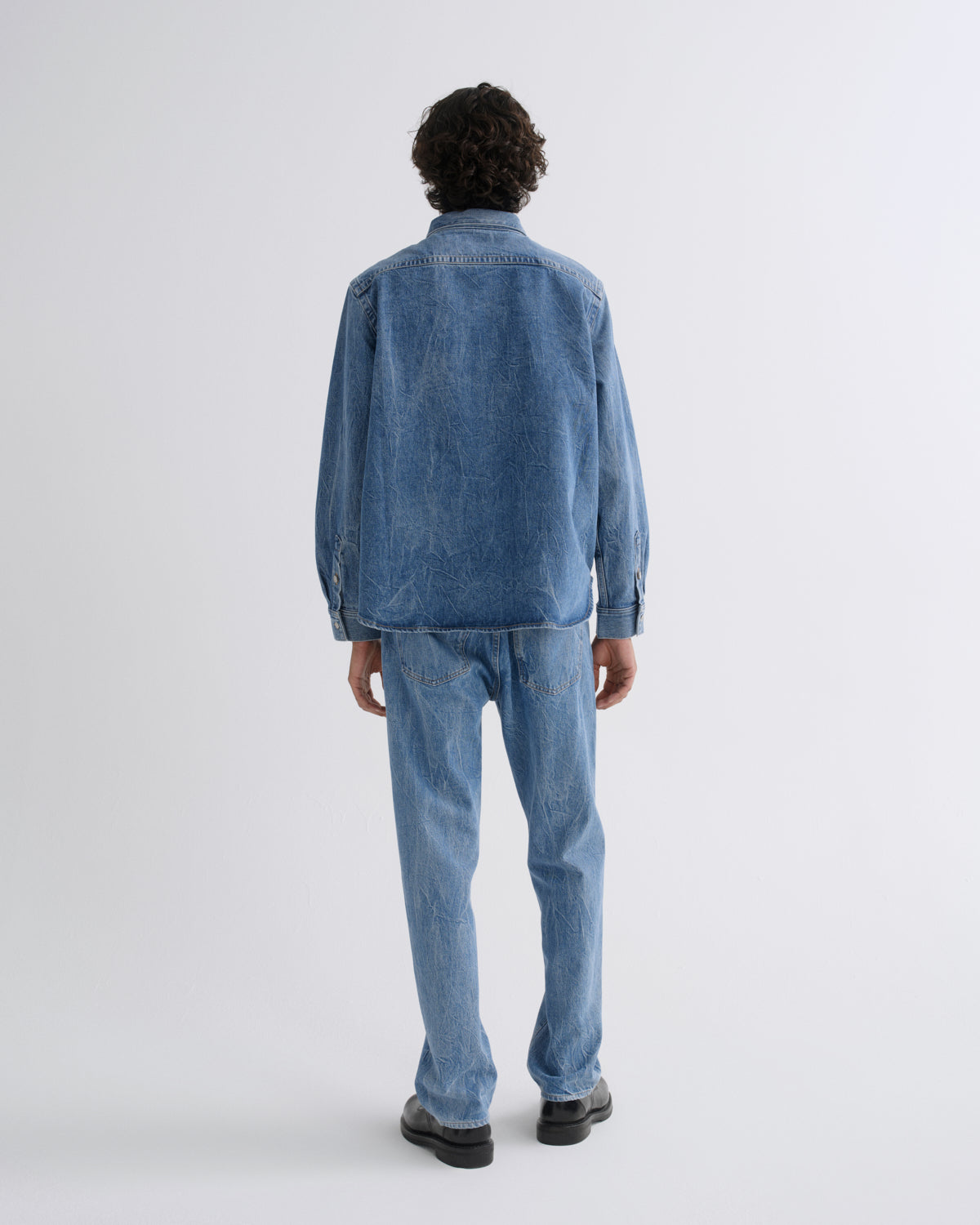 A model viewed from the back wearing a blue denim jacket and matching blue denim jeans, standing against a plain light gray studio background.
