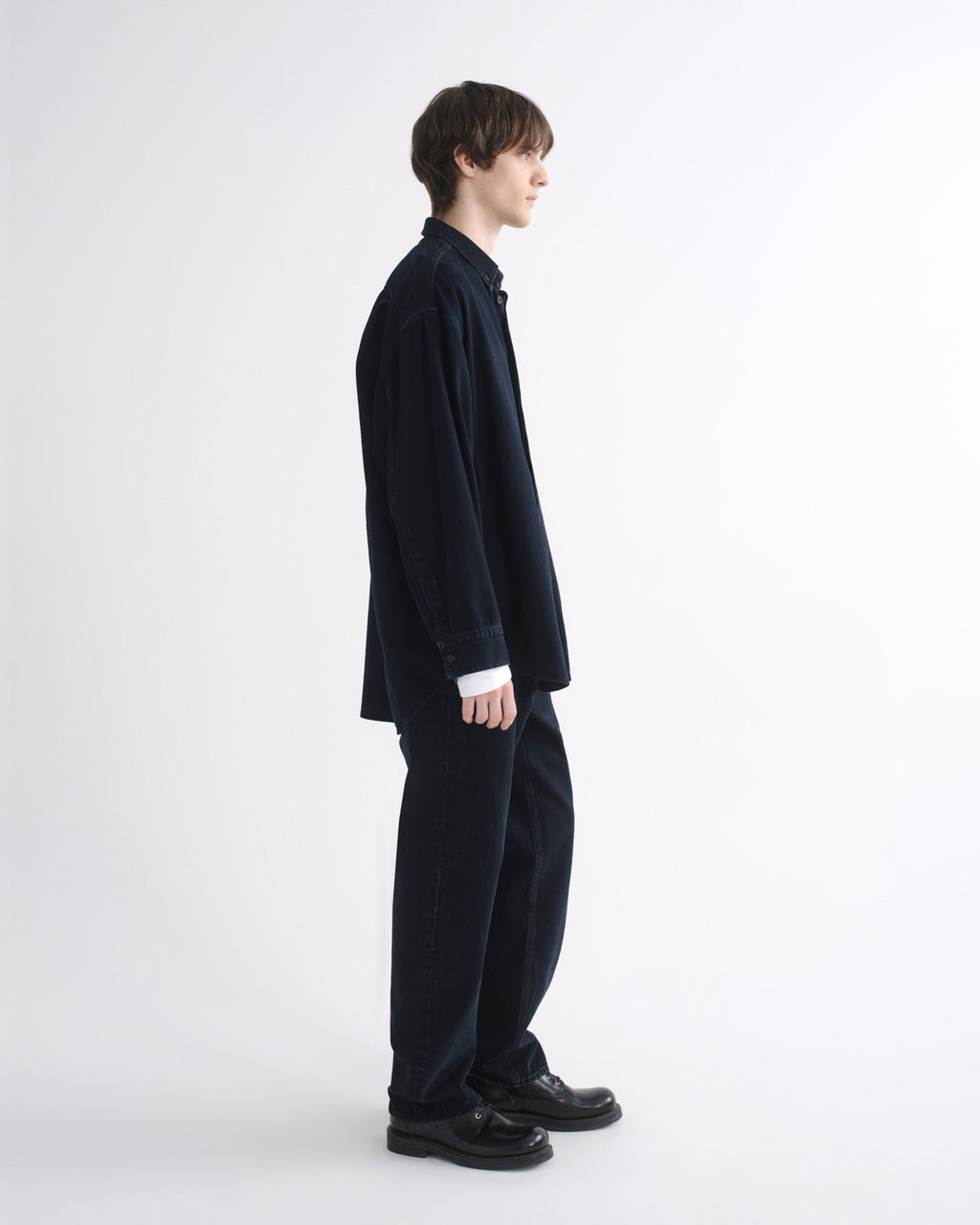 Person wearing a black long sleeve shirt with black pants and black shoes, side view, on a grey background