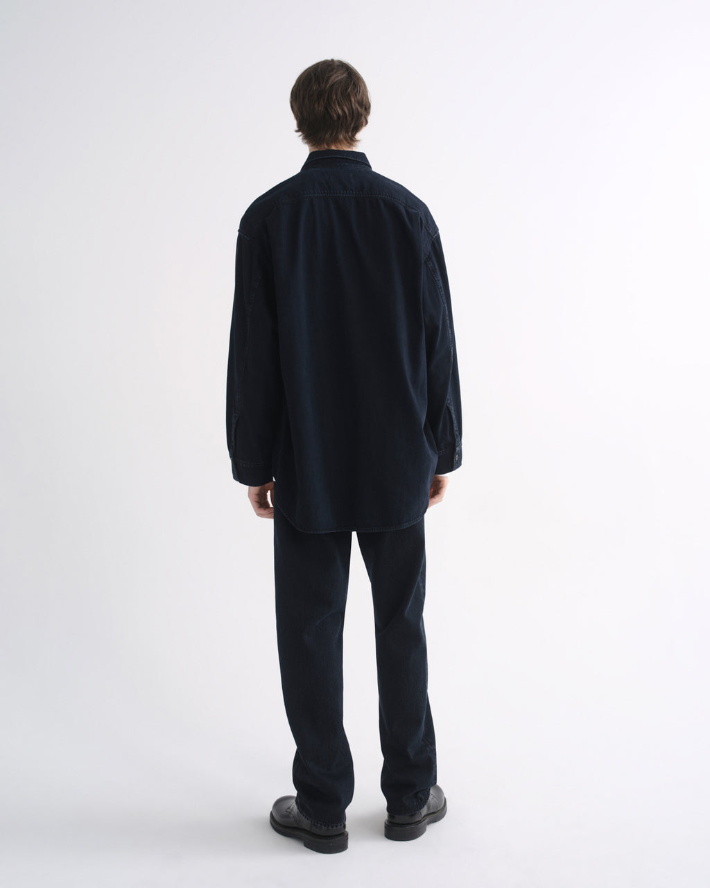 Person wearing a black long sleeve shirt with black pants and black shoes, back view, on a grey background