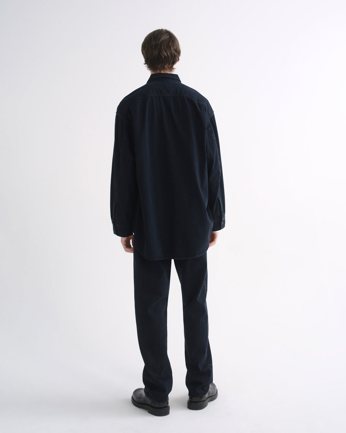 Person wearing a black long sleeve shirt with black pants and black shoes, back view, on a grey background