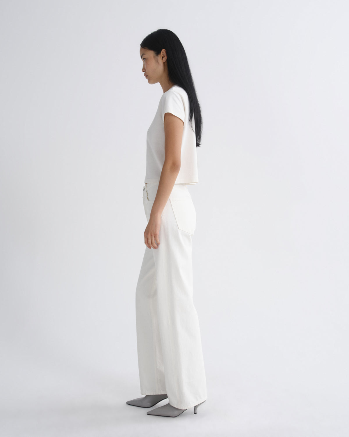A woman in a white short-sleeve crop top and white wide-leg trousers stands in profile against a plain light gray studio background, wearing gray pointed-toe heels.