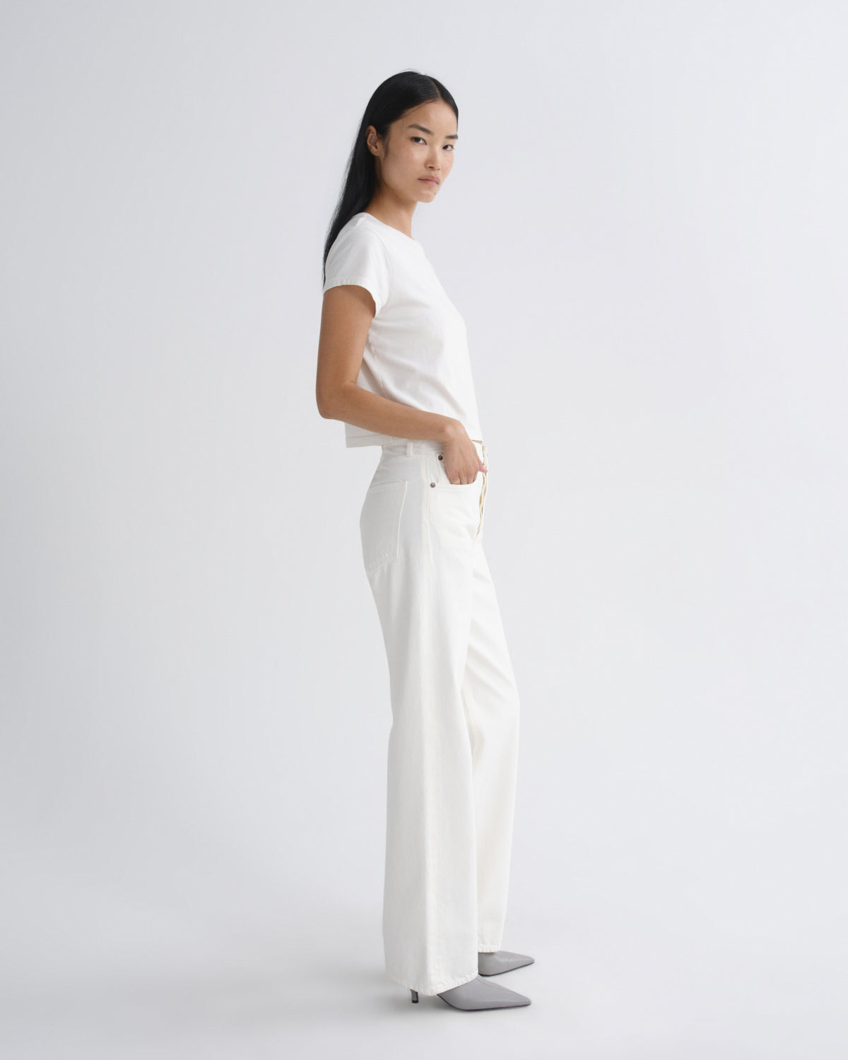 A woman modeling a white short-sleeve crop top and matching white wide-leg trousers, standing in profile against a clean light gray studio background.