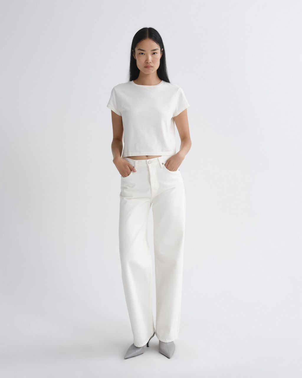 A model stands against a light gray studio background wearing a white short-sleeve cropped T-shirt, white wide-leg pants, and gray pointed shoes.