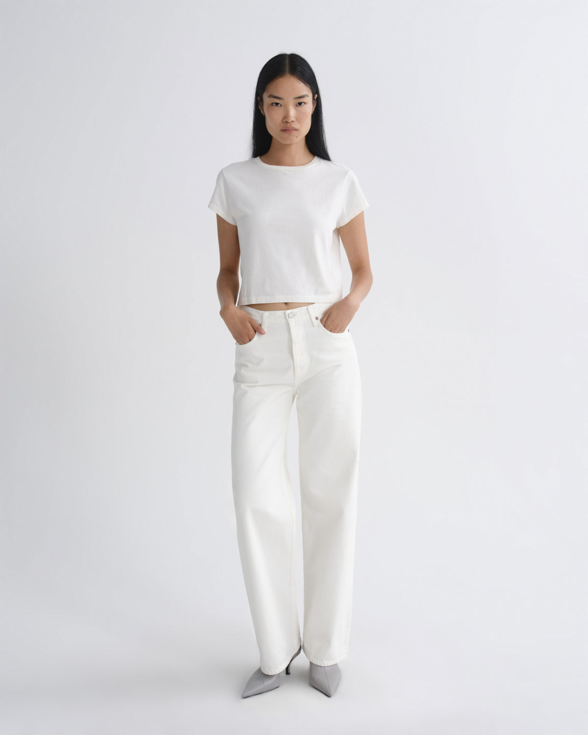 A model stands against a light gray studio background wearing a white short-sleeve cropped T-shirt, white wide-leg pants, and gray pointed shoes.