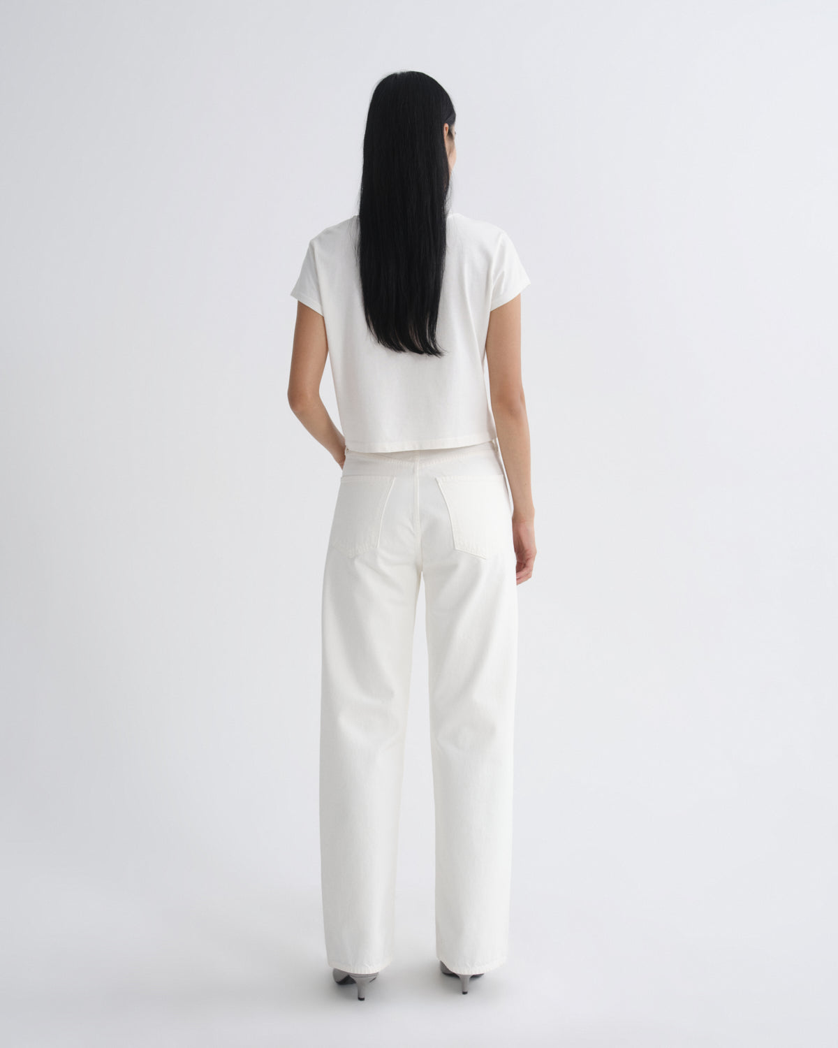 A model viewed from the back wearing a white short-sleeve cropped top and white wide-leg trousers against a plain light gray studio background.
