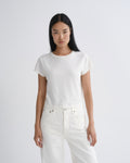 A model with long black hair is wearing a white short-sleeve crewneck t-shirt and white jeans, standing against a light gray studio background.