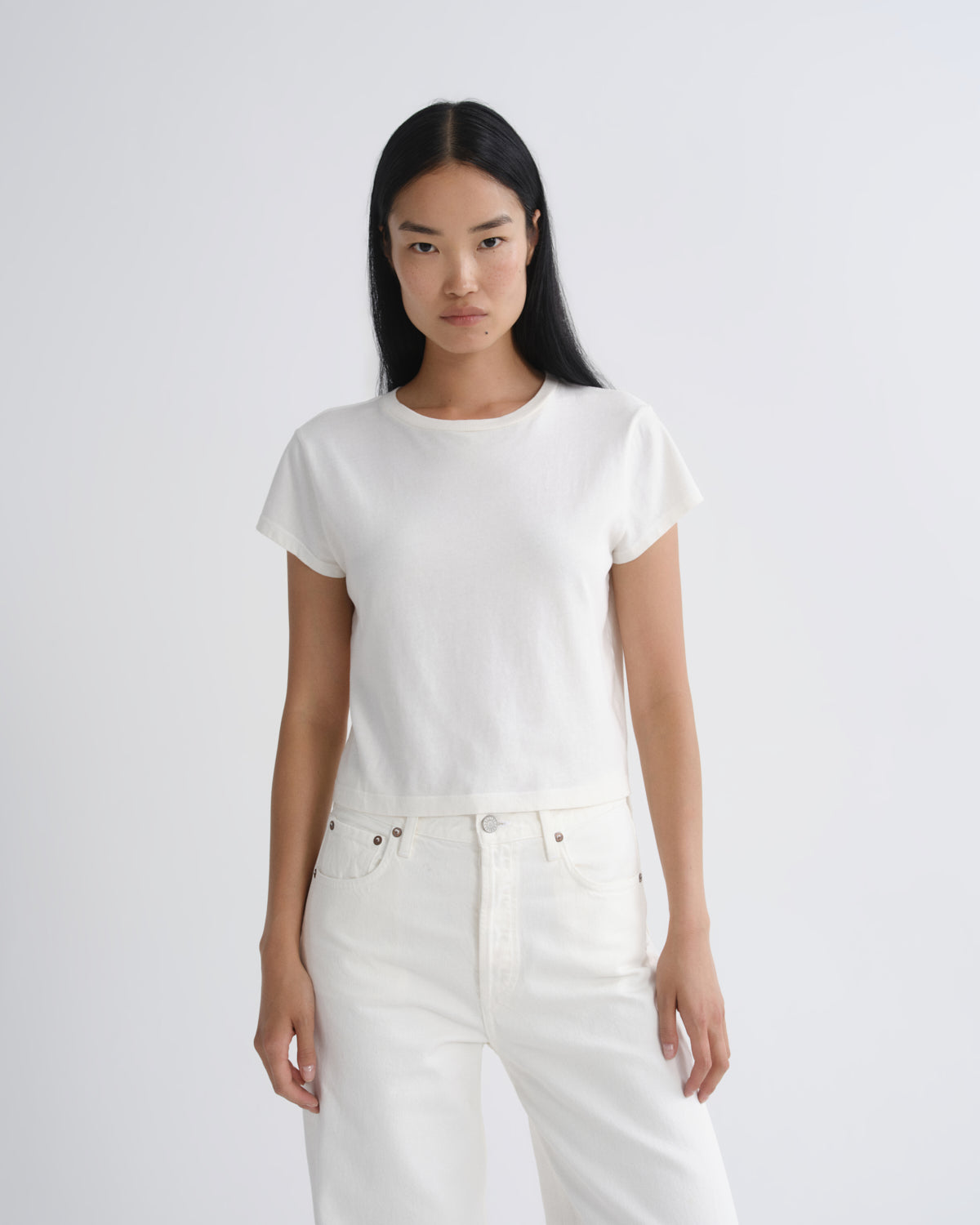 A model with long black hair is wearing a white short-sleeve crewneck t-shirt and white jeans, standing against a light gray studio background.