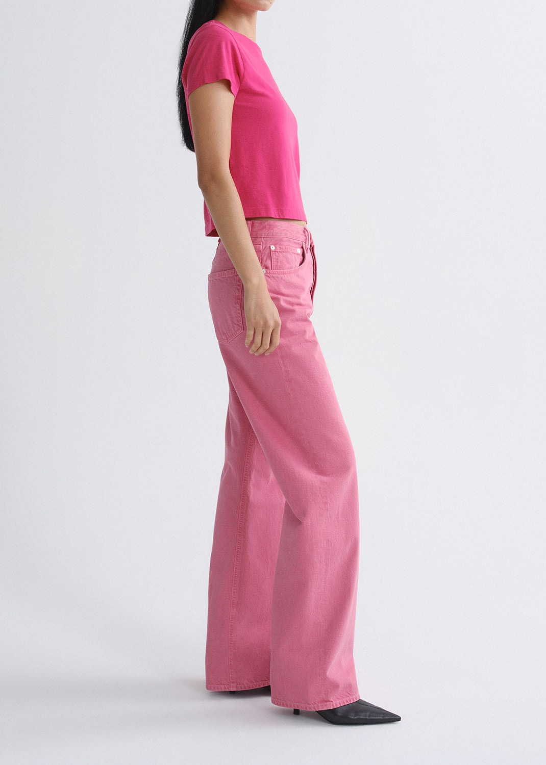 Person wearing a bright pink short sleeve crewneck tee with light pink straight leg jeans viewed from the side on a grey background
