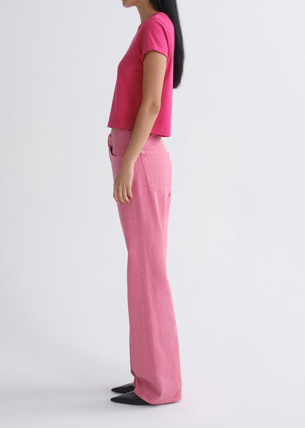 Person wearing a bright pink short sleeve crewneck tee with light pink straight leg jeans viewed from the side on a grey background
